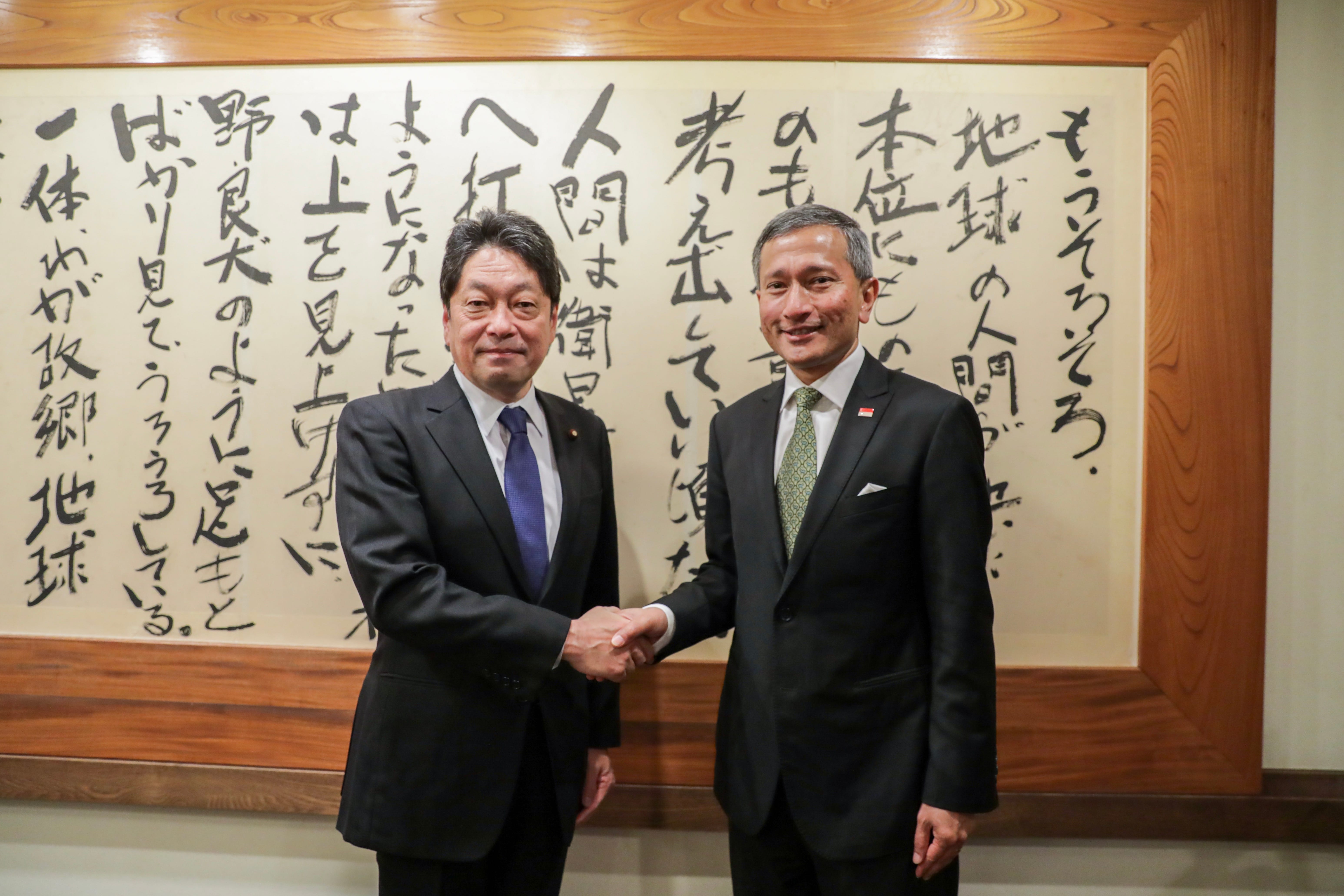Two men in suits shaking hands, standing in front of a framed calligraphy scroll.